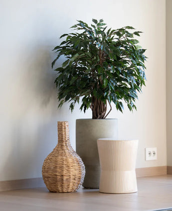 Een Kunstplant Ficus Liana 90 cm staat in een grijze plantenbak naast een gevlochten vaas en een licht geribbeld krukje, alles op een houten vloer tegen een effen witte muur.