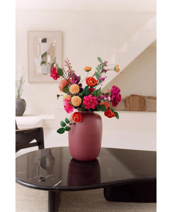De Vaas Artic Roze Ø21 x H29 cm, gevuld met roze, oranje en rode bloemen, staat op een donker glanzende salontafel in een moderne minimalistische woonkamer met een trap en ingelijste kunstwerken op de achtergrond.