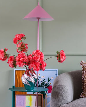 Een roze lamp, een gestreepte vaas met het Zijden kunstbloemenboeket Kersenbloesem Koraal, kleurrijke kunstwerken en een grijze stoel staan tegen een lichtgroene muur, waardoor een gezellige, levendige hoek ontstaat.