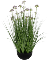 De Kunstgras Prei Bloem Ui Graspot 60cm brandvertragend heeft hoge, dunne groene grasachtige bladeren en delicate kleine paarse bloemen in een zwarte pot, tegen een witte achtergrond.